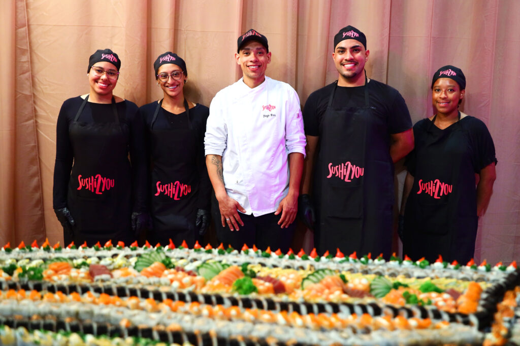 Cinco funcionários da Sushi 2 You, vestindo uniformes pretos e gorros com o logotipo da empresa, sorrindo para a câmera, atrás de uma extensa mesa coberta com várias fileiras de sushis e sashimis. Um dos funcionários veste um dólmã branco de chef.